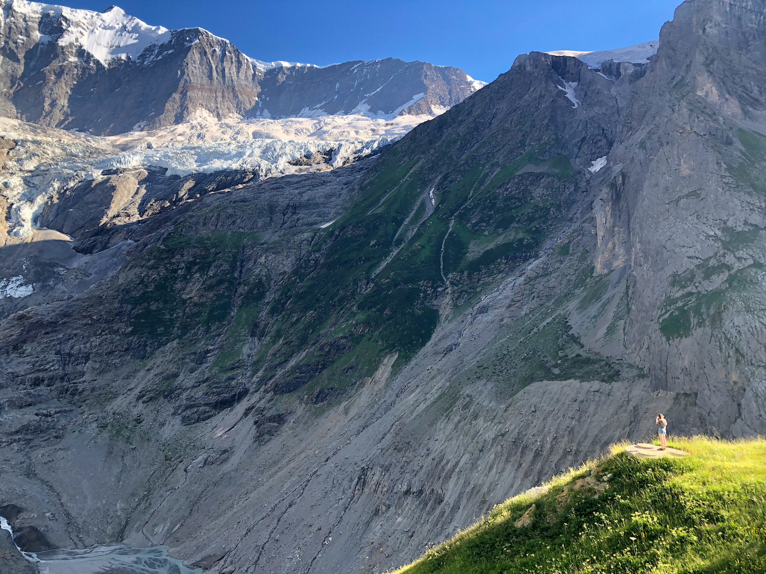 image: A person stands on a grassy hill observing mountains with a retreated glacier.