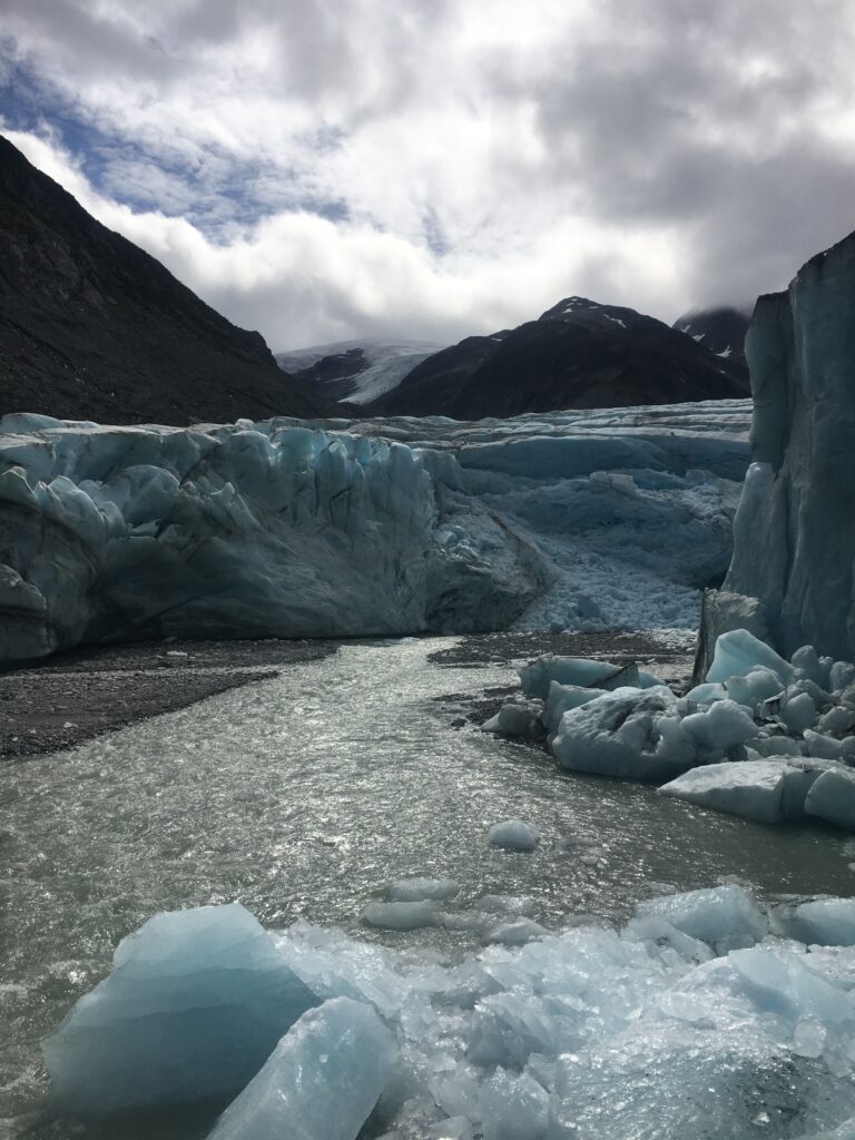 A glacier with a foreground of icy, fragmented waters, set against a backdrop of mountains under a cloudy sky
