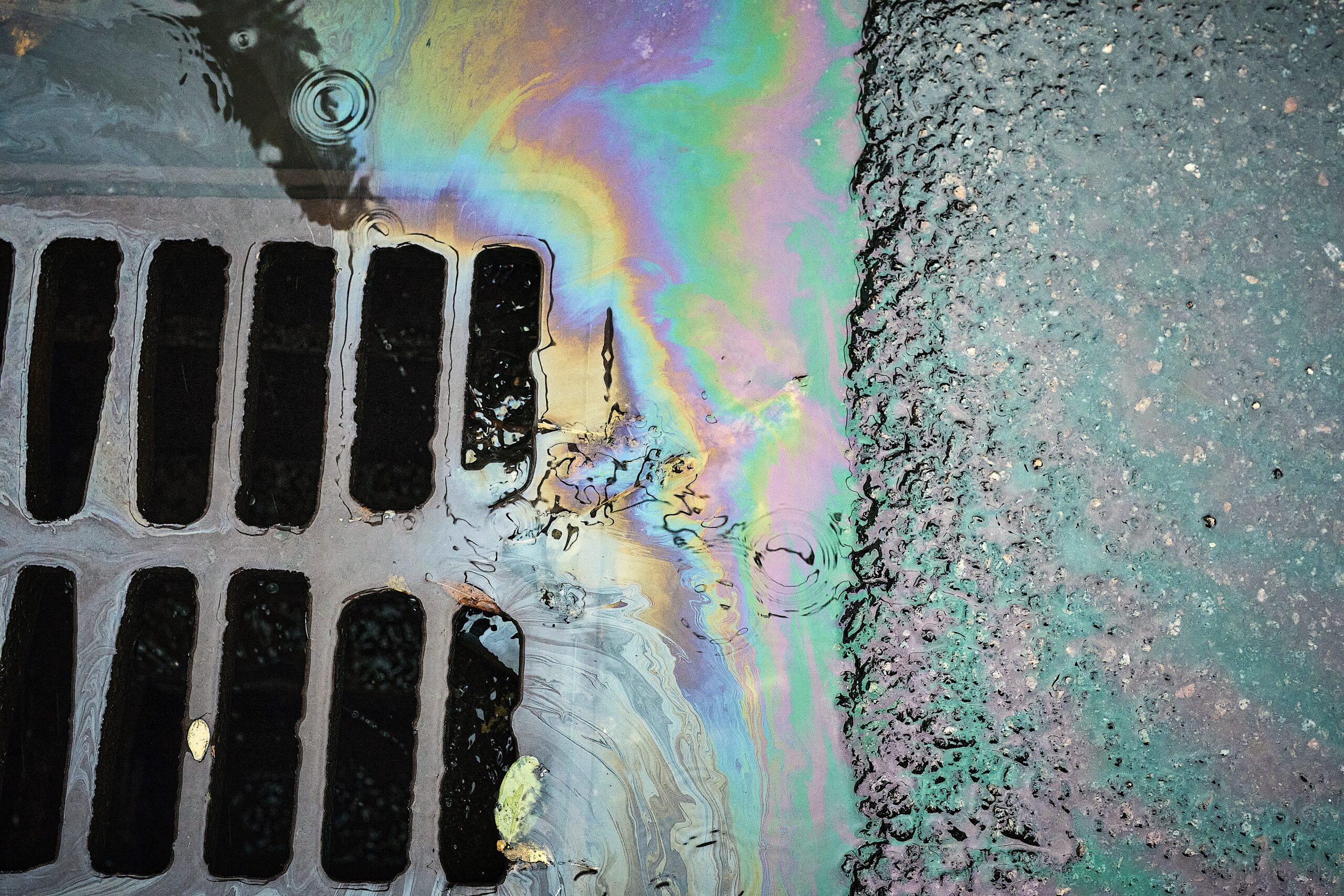image: Colorful Oil Sheen Reflecting in Water Near a Storm Drain on a Rainy Day in an Urban Environment.