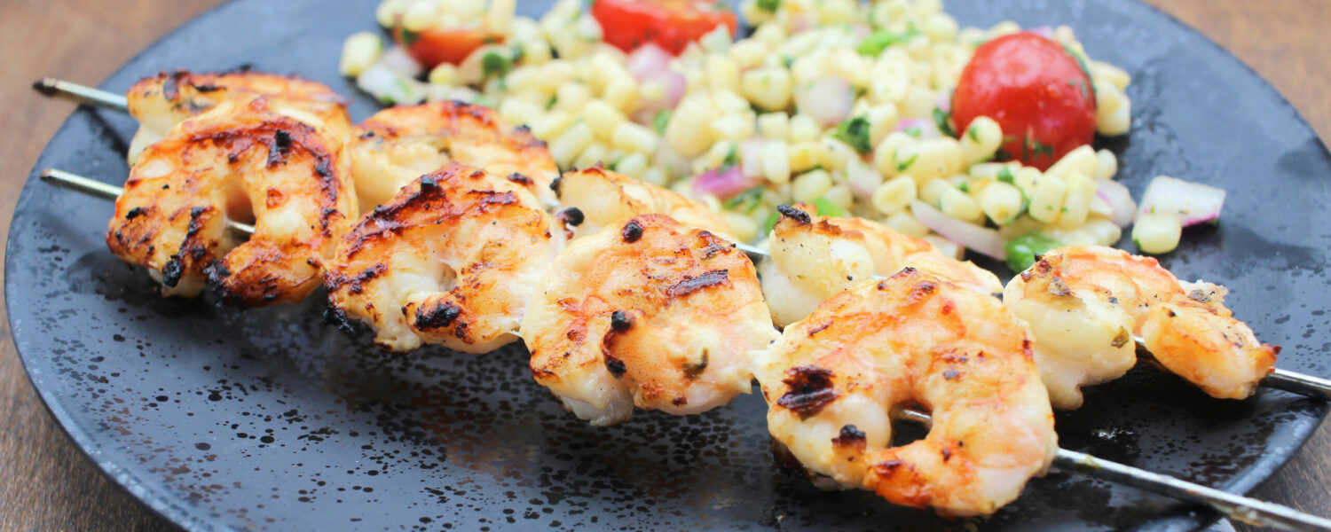image: shrimp on skewers on a plate with tomatoes and corn.