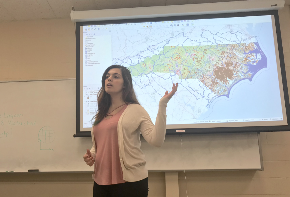 image: Emine Fidan speaking in a classroom in front of a projector with a photo of the map of North Carolina. Credit: NC State University's Department of Biological and Agricultural Engineering.