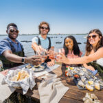Four people enjoy NC oysters and wine with Stump Sound, N.C., in the background. Photo by Justin Kase Conder