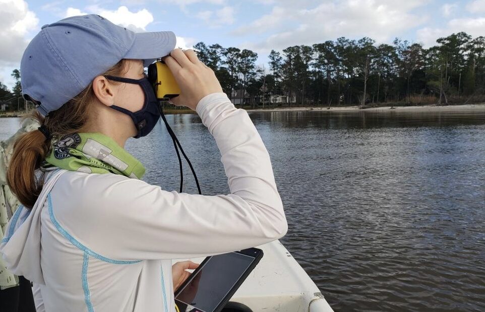 A girl in a blue hat and white shirt looks through binoculars while aboard a small boat