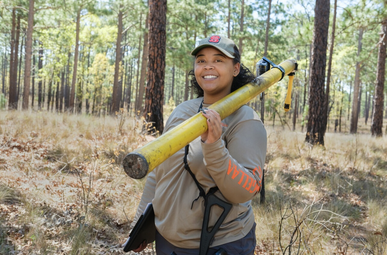 image: Lauren smiles while holding a clipboard, a pole with a camera, and a spotting scope.