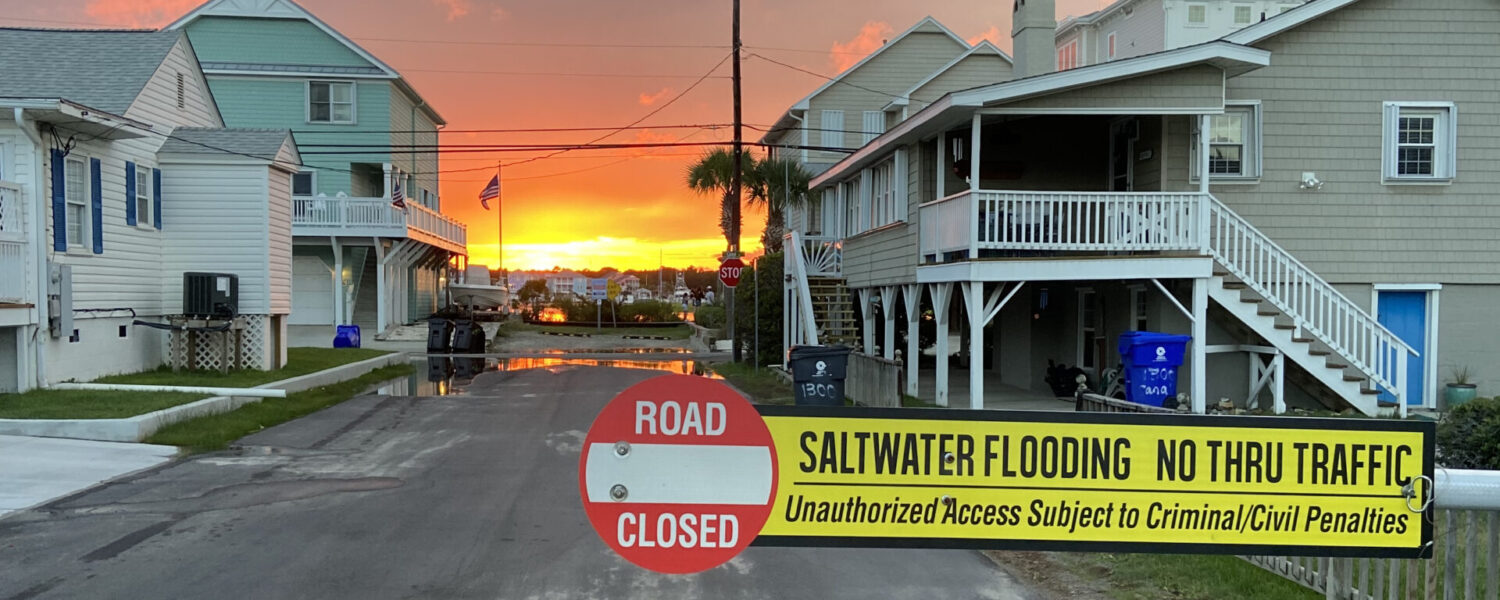 Flooding is occurring at a residential intersection in the background, and a sign is present in the foreground that reads, "Road Closed. Saltwater Flooding. No Thru Traffic. Unauthorized Access Subject to Criminal/Civil Penalties. Photo credit: Natalie Nelson