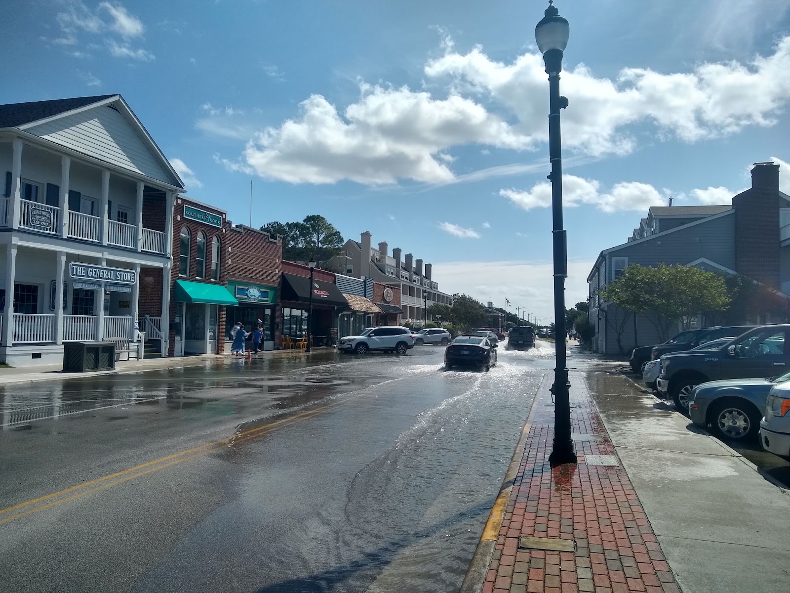 An urban street is partly flooded with saltwater from a high tide flood. Cars are parked in and driving through the floodwaters.