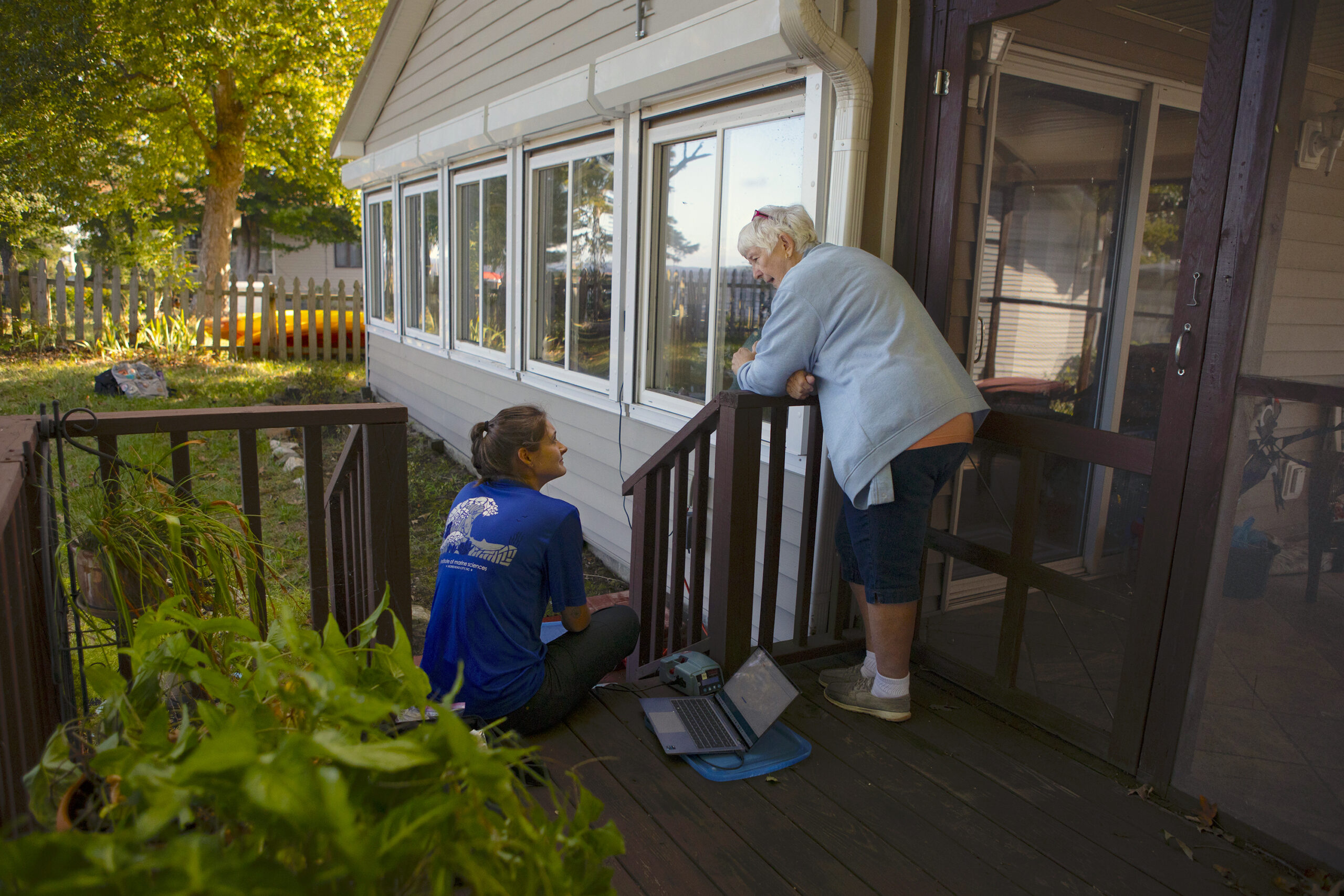 Local community member leans over the deck balcony while talking to a researcher, seated below.