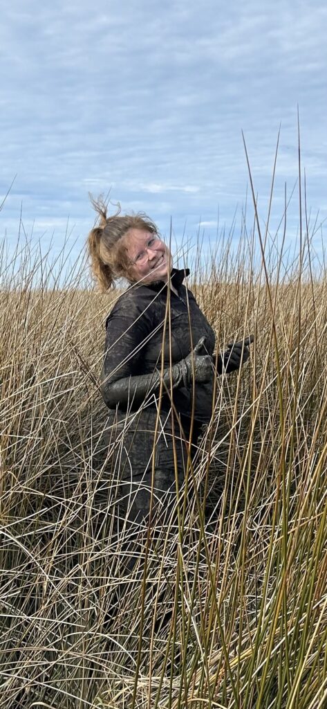 A woman standing in tall grasses doing two thumbs ups