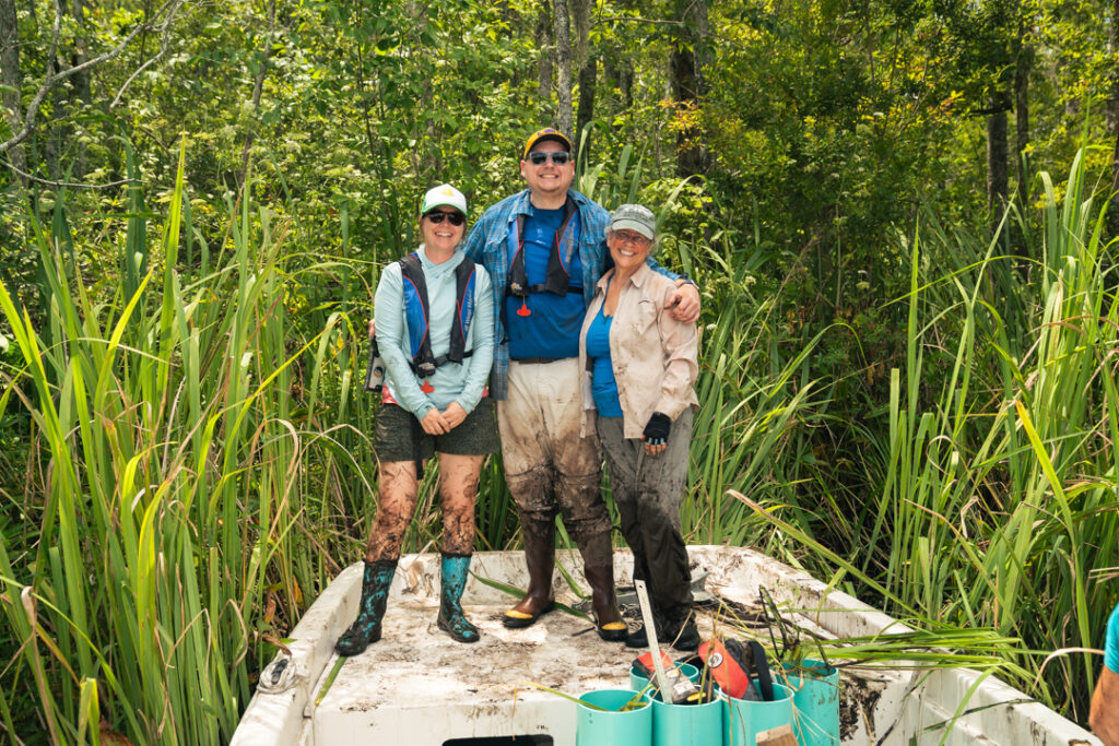 Two women and a man standing at the end of a boat in a wetland, everyone covered in mud