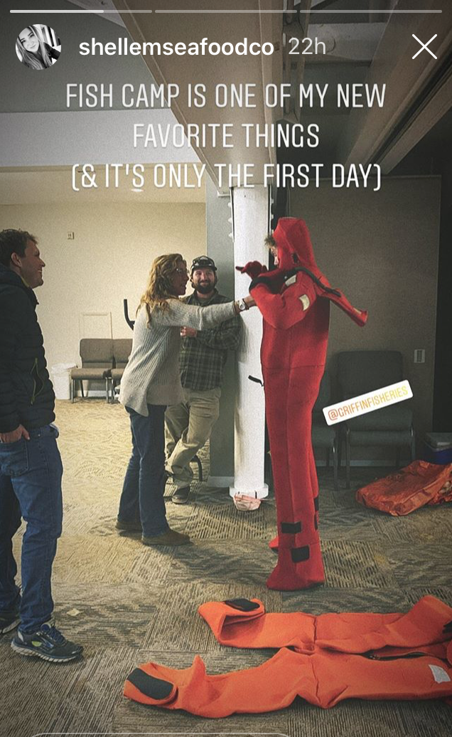 Four people indoors during a Fish Camp training session, with one person fully suited in a red survival (immersion) suit while others look on, smiling and offering assistance. Another red suit lies on the floor in the foreground. Text over the image reads: