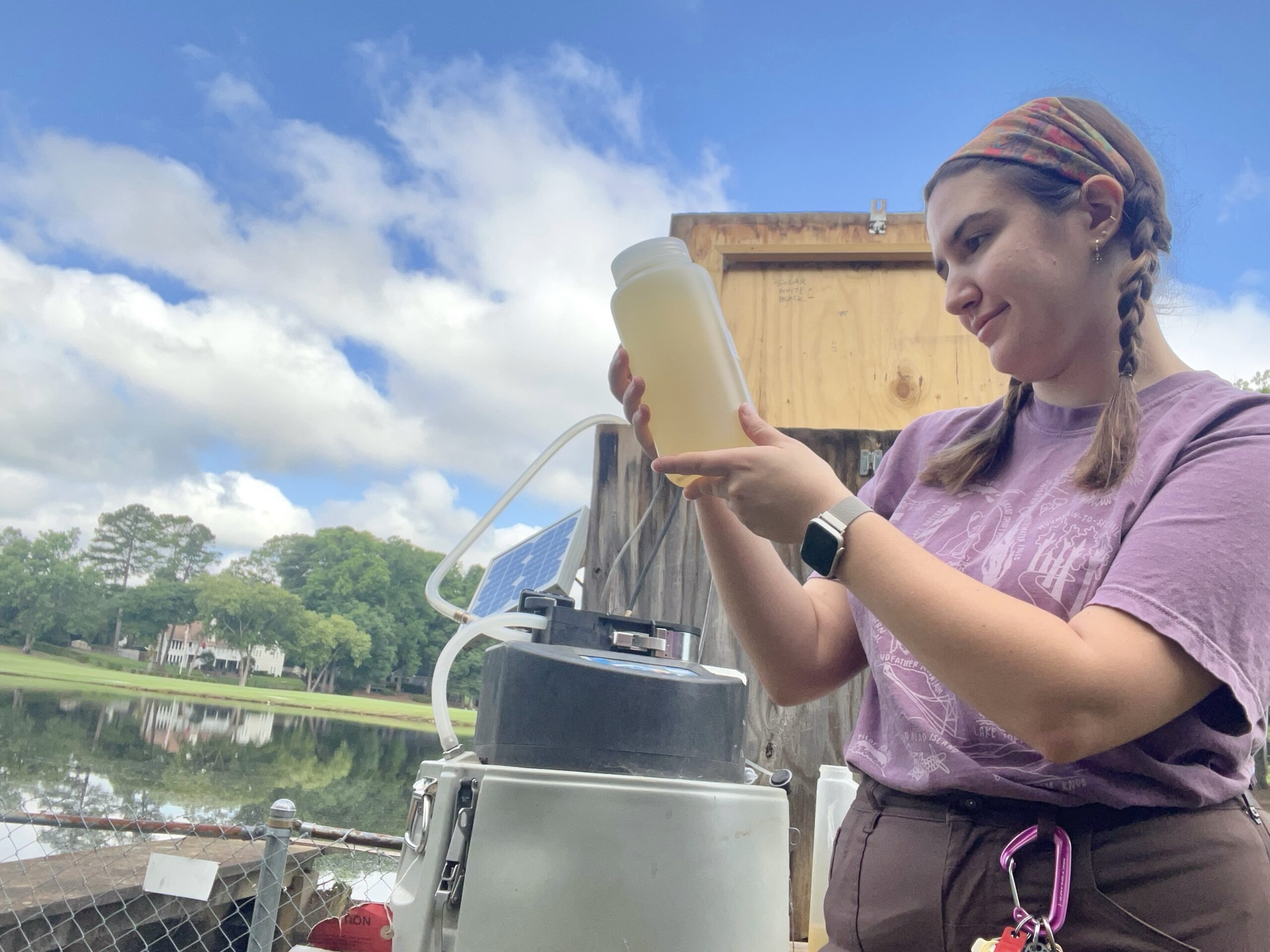 A woman holding a bottle-like container with water in it by a pond