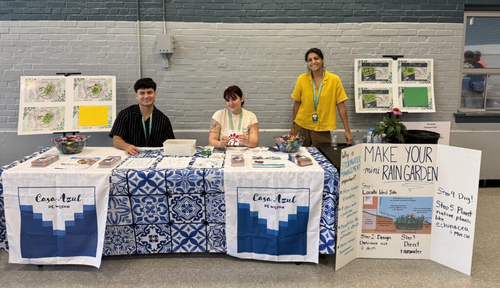 Two adults sit and one adult stands behind tables with Casa Azul de Wilson signs and a "Make Your Mini Rain Garden" poster