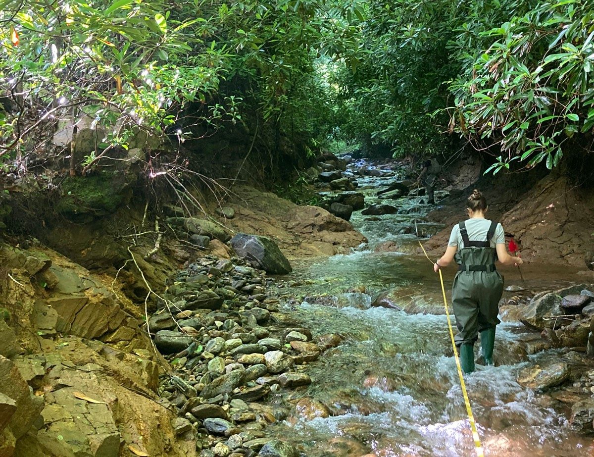 A person in waders walks through a rocky creek holding onto a rope line